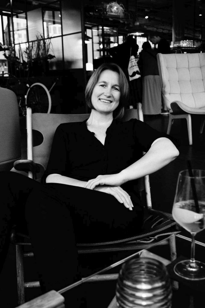 Yvonne Griesel, entspannt lächelnd in einem Restaurantstuhl mit Glas auf einem Tisch vor sich