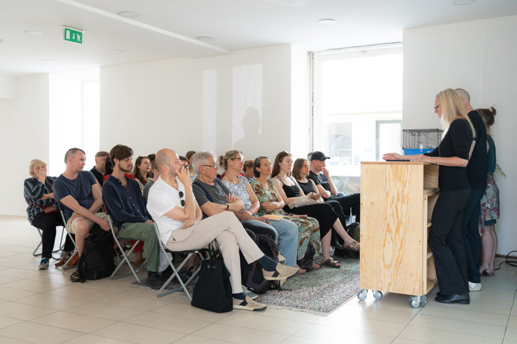 Eine Gruppe von Menschen sitzt in einem modernen Raum während einer Veranstaltung. Im Vordergrund steht eine Frau am Rednerpult und spricht zu den Anwesenden. Die Teilnehmer scheinen aufmerksam zuzuhören.