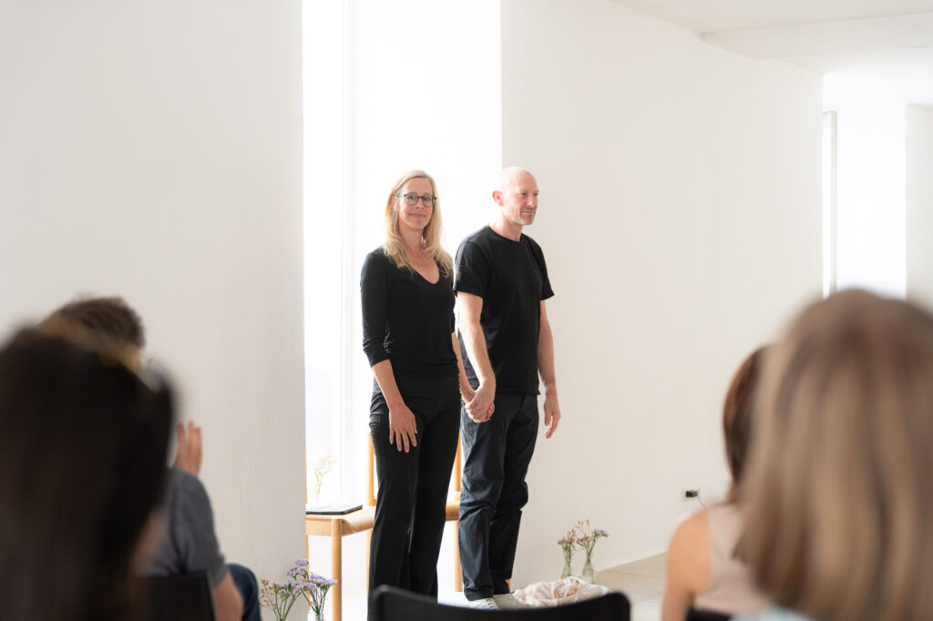 Zwei Personen stehen nebeneinander auf einer Bühne, während das Publikum ihnen zuhört. Die Umgebung ist hell und modern, mit Pflanzen auf der Bühne.